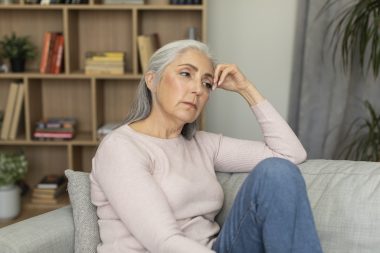 Sad tired depressed caucasian senior woman sitting on sofa, suffering from menopause, migraine