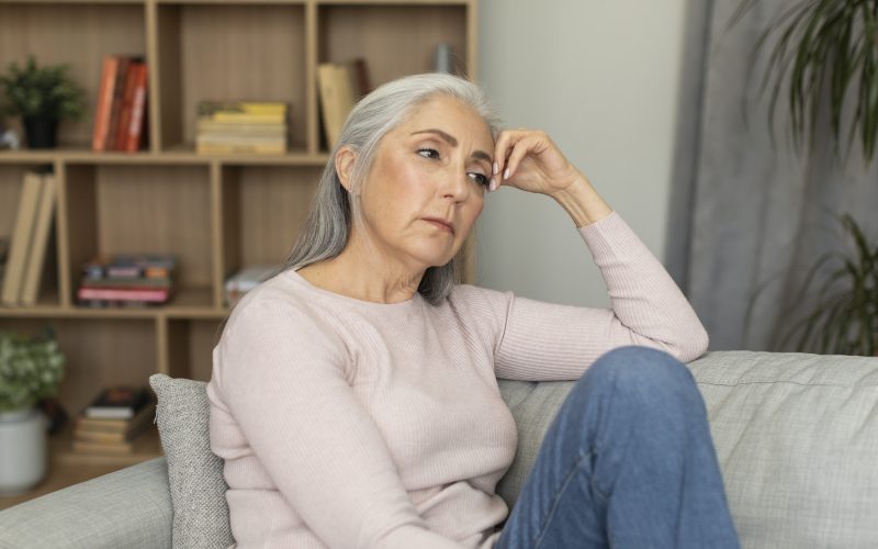 Sad tired depressed caucasian senior woman sitting on sofa, suffering from menopause, migraine